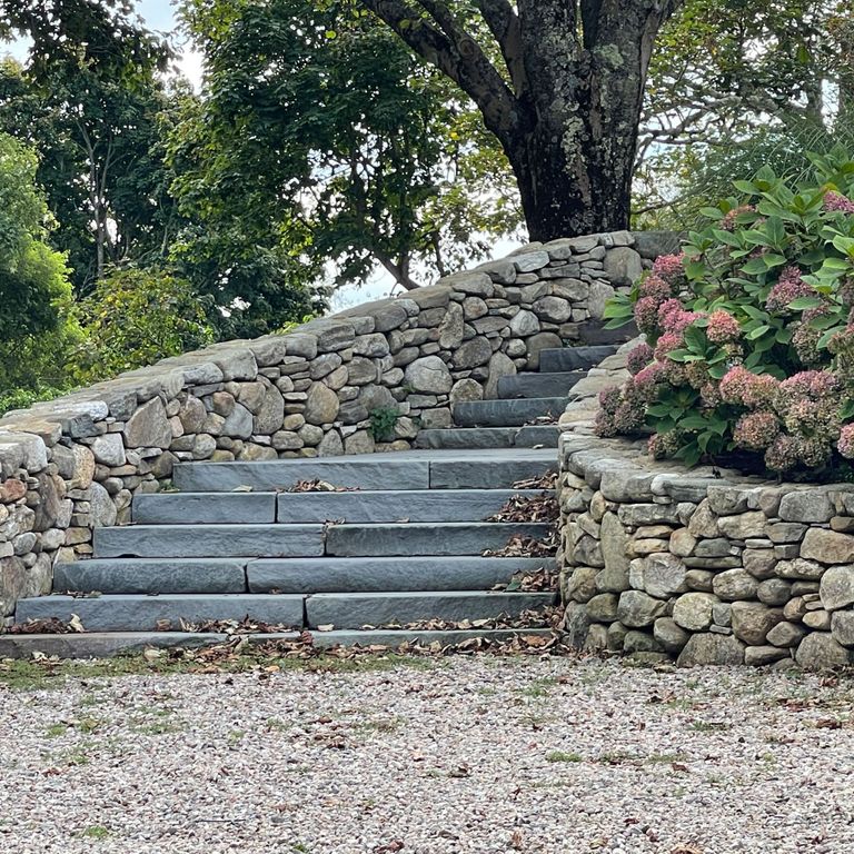 Feature image for New England Fieldstone Mix. The image shows this material being used in a landscape wallstone application. The surface finish shown is weathered. The dominant color shown is gray; with brown, green and tan secondaries. The color mix creates a warm feeling.