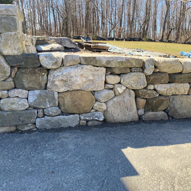 Application image for New England Fieldstone Mix. The image shows this material being used in a landscape wallstone application. The surface finish shown is weathered. The dominant color shown is gray; with brown, green and tan secondaries. The color mix creates a cool feeling. Regular, blotched and random color patterns.