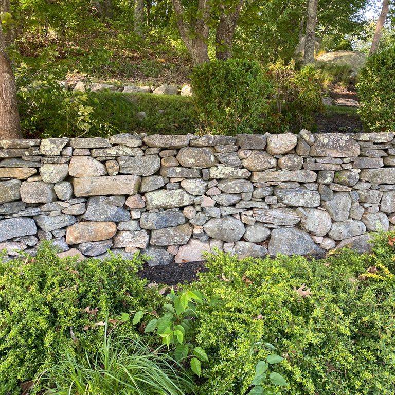 Application image for New England Fieldstone Mix. The image shows this material being used in a landscape wallstone application. The surface finish shown is weathered. The dominant color shown is gray; with beige, brown, white, tan and cream secondaries.