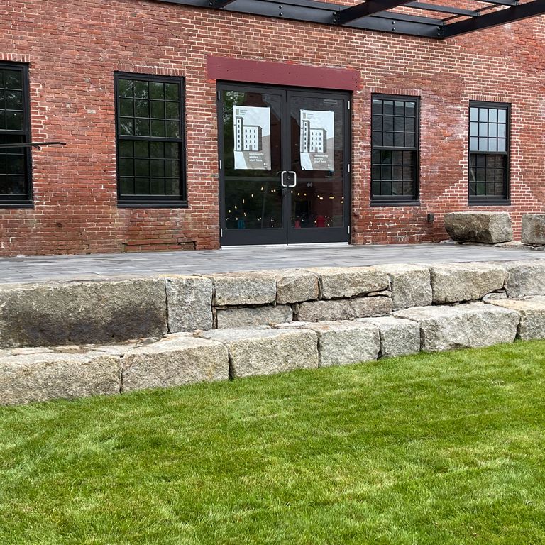 Application image for Reclaimed Granite Block. The image shows this material being used for landscape wallstone and monolithic steps. The surface finish shown is a combination of split-face and weathered. Dominant colors shown are beige, gray and tan; with brown and cream secondaries. The color mix creates a warm feeling. Speckled color pattern.
