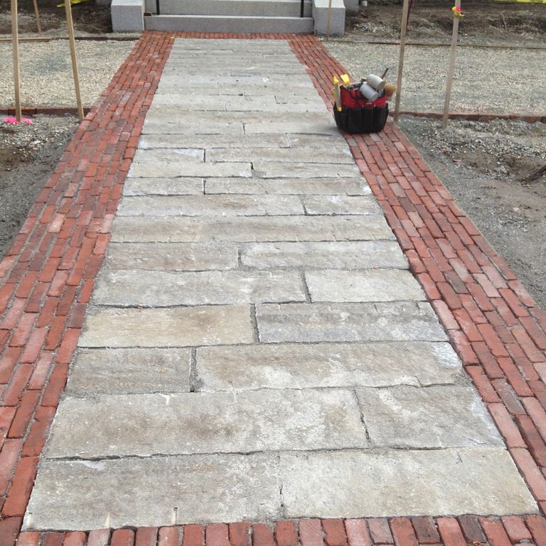 Feature image for Reclaimed Granite Curbstone - Junior. The image shows this material being used in a path pavers application. The design pattern shown is roughly square roughly rectangular. The dominant color shown is gray; with beige, white, tan and cream secondaries. Regular and blotched color patterns.