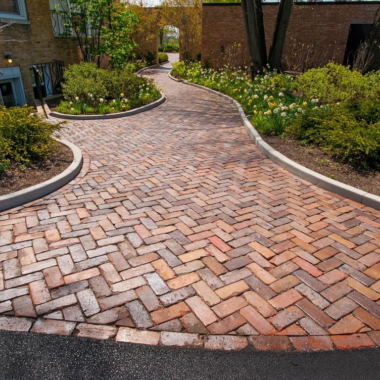 Feature image for Reclaimed Midwest Brick Pavers. The image shows this material being used for driveway pavers and path pavers. The design pattern shown is roughly square roughly rectangular. Dominant colors shown are brown and orange/rust; with beige, gray, pink, purple and tan secondaries. The color mix creates a warm feeling. Regular color pattern.