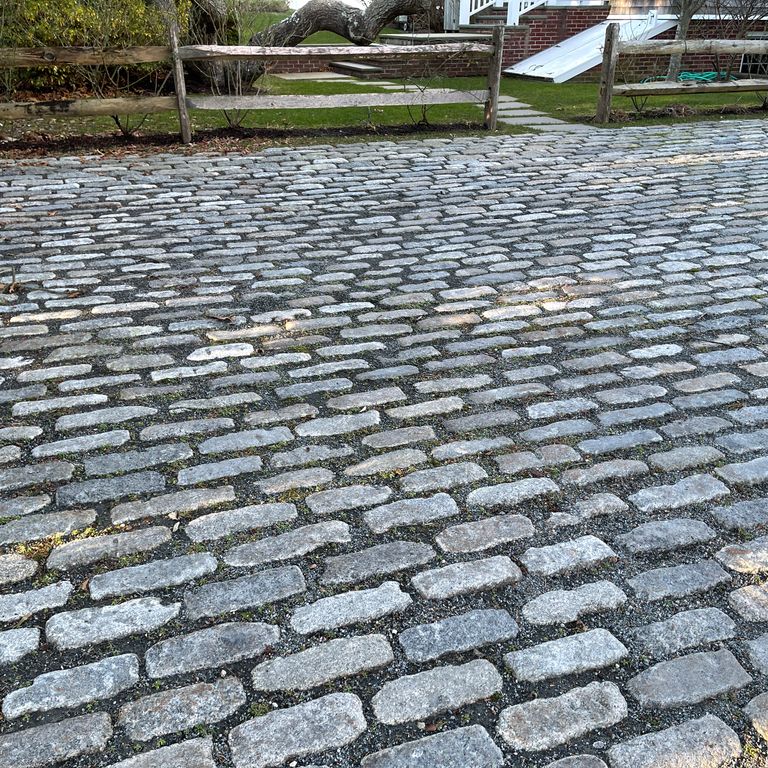 Feature image for Reclaimed New England Granite Cobblestones - Jumbo. The image shows this material being used in a driveway pavers application. Design patterns shown are dimensional squares and rectangles, random with wide sides up and random with tops up. Dominant colors shown are blue and gray; with green, purple, white and tan secondaries. The color mix creates a cool feeling. Regular and speckled color patterns. Contains some mica and mineral inclusions.