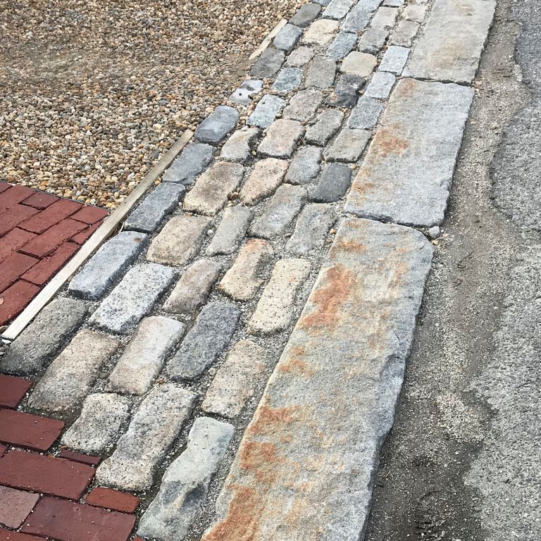 Application image for Reclaimed New England Granite Cobblestones - Jumbo. The image shows this material being used in a driveway aprons application. The surface finish shown is a combination of weathered and worn. The dominant color shown is gray; with beige, brown, green, pink, purple, white, tan and cream secondaries. The color mix creates a warm feeling. Speckled and blotched color patterns.