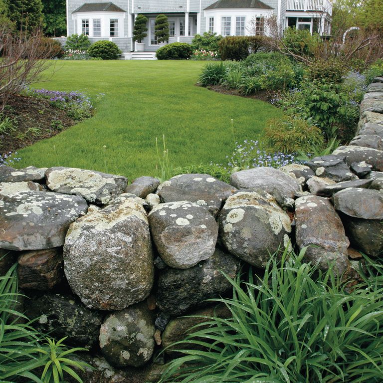 Feature image for New England Fieldstone Rounds. The image shows this material being used in a landscape wallstone application. The surface finish shown is weathered. The dominant color shown is gray; with blue, brown and white secondaries. The color mix creates a cool feeling. Fine speckled, blotched and random color patterns.