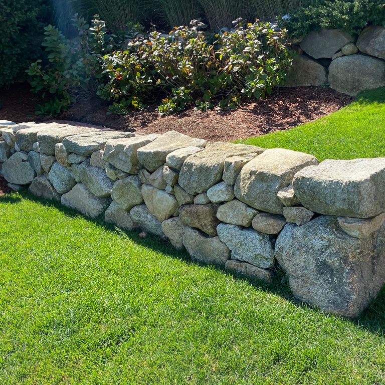 Application image for New England Fieldstone Rounds. The image shows this material being used in a landscape wallstone application. The surface finish shown is weathered. The dominant color shown is gray.
