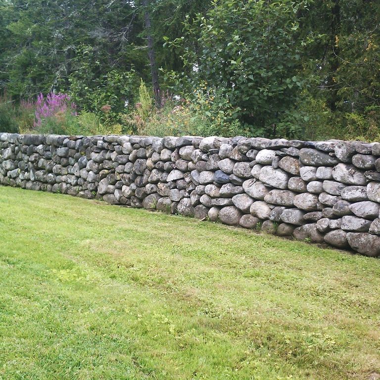 Application image for New England Fieldstone Rounds. The image shows this material being used in a landscape wallstone application. The surface finish shown is weathered. The dominant color shown is gray.