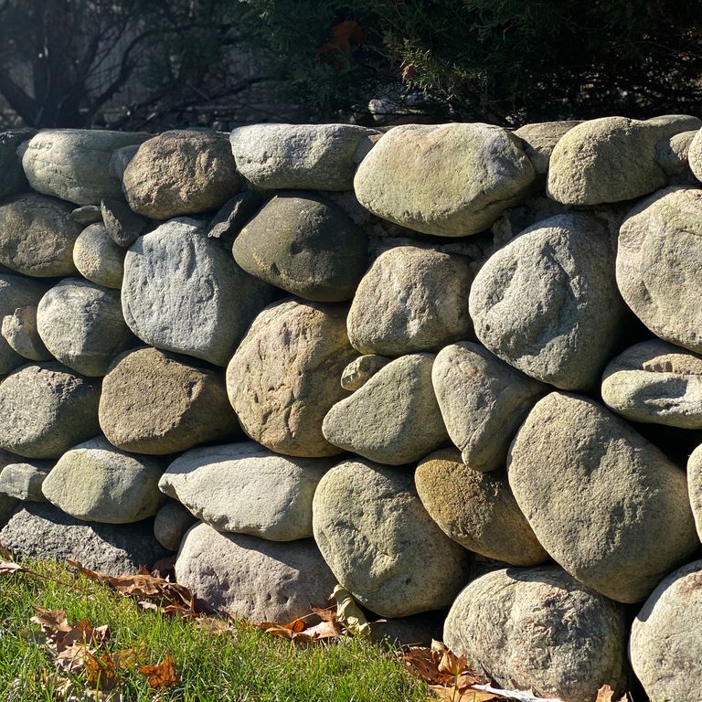 Application image for New England Fieldstone Rounds. The image shows this material being used in a landscape wallstone application. The surface finish shown is weathered. The dominant color shown is gray.