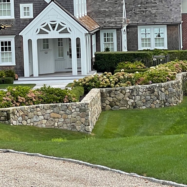 Application image for New England Fieldstone Rounds. The image shows this material being used in a landscape wallstone application. The surface finish shown is weathered. The dominant color shown is gray; with beige, brown and tan secondaries. The color mix creates a warm feeling.