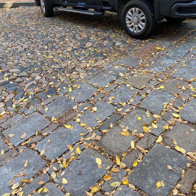 Feature image for Reclaimed New England Granite Cobblestones - Road Pavers. The image shows this material being used in a driveway pavers application. The design pattern shown is dimensional squares and rectangles. The dominant color shown is gray; with black, brown, orange/rust, purple and tan secondaries. Speckled color pattern.