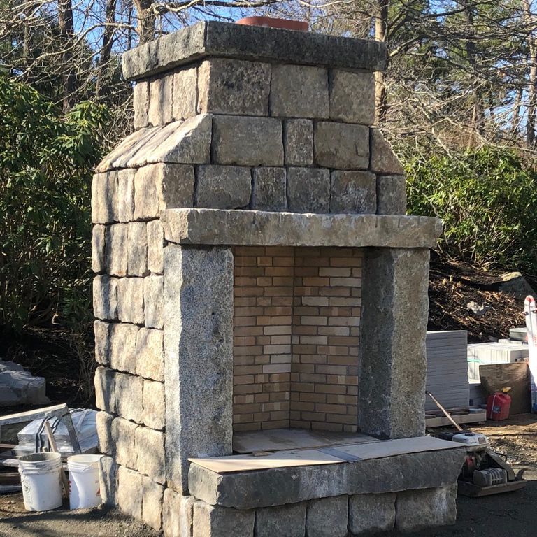Application image for Reclaimed New England Granite Cobblestones - Road Pavers. The image shows this material being used for fireplace surround and outdoor fire concepts. The surface finish shown is a combination of weathered and worn. Dominant colors shown are beige and tan; with brown, gray, orange/rust, white, yellow and cream secondaries.