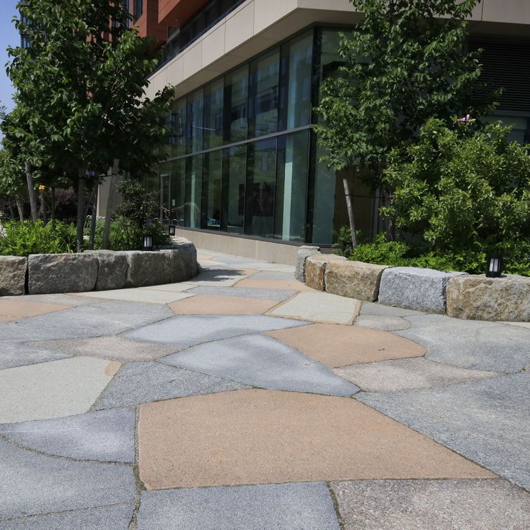 Application image for Granite - New England Mix. The image shows this material being used for patio pavers and public area. The surface finish shown is flamed. Dominant colors shown are beige and gray. Fine speckled and speckled color patterns.