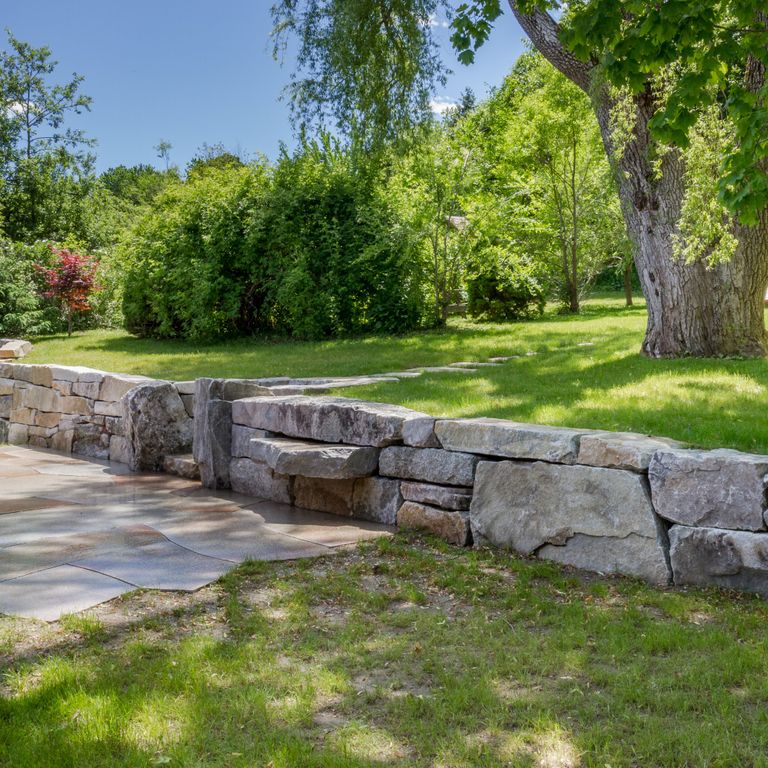 Feature image for Black Mountain Granite - Natural. The image shows this material being used for landscape wallstone and patio pavers. The design pattern shown is quarried-snapped random ashlar. The dominant color shown is gray; with beige, blue, brown, orange/rust and tan secondaries. Random, speckled and blotched color patterns.
