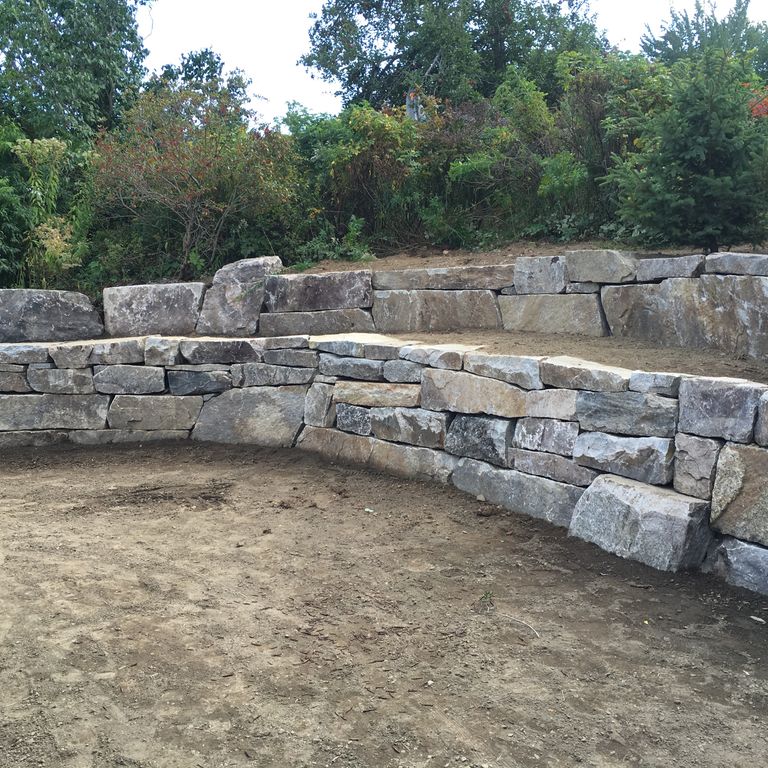 Application image for Black Mountain Granite - Natural. The image shows this material being used in a landscape wallstone application. The surface finish shown is split-face. The dominant color shown is gray; with black, blue, brown, green, purple and white secondaries. Regular color pattern.