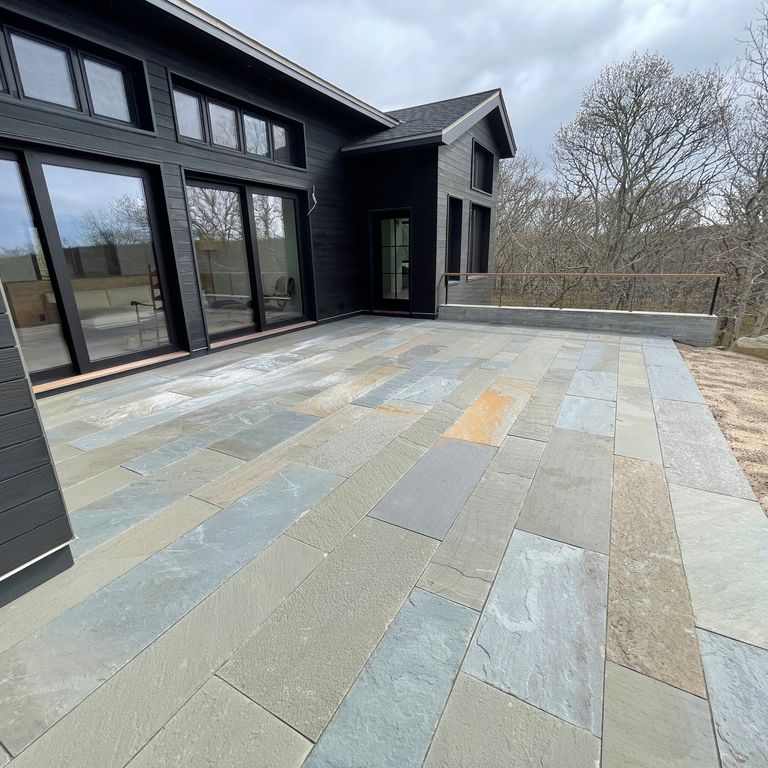 Feature image for Bluestone Natural Cleft - Full Color Mix. The image shows this material being used in a patio pavers application. The surface finish shown is natural cleft. The dominant color shown is gray; with blue and orange/rust secondaries. The color mix creates a cool feeling. Regular and random color patterns.