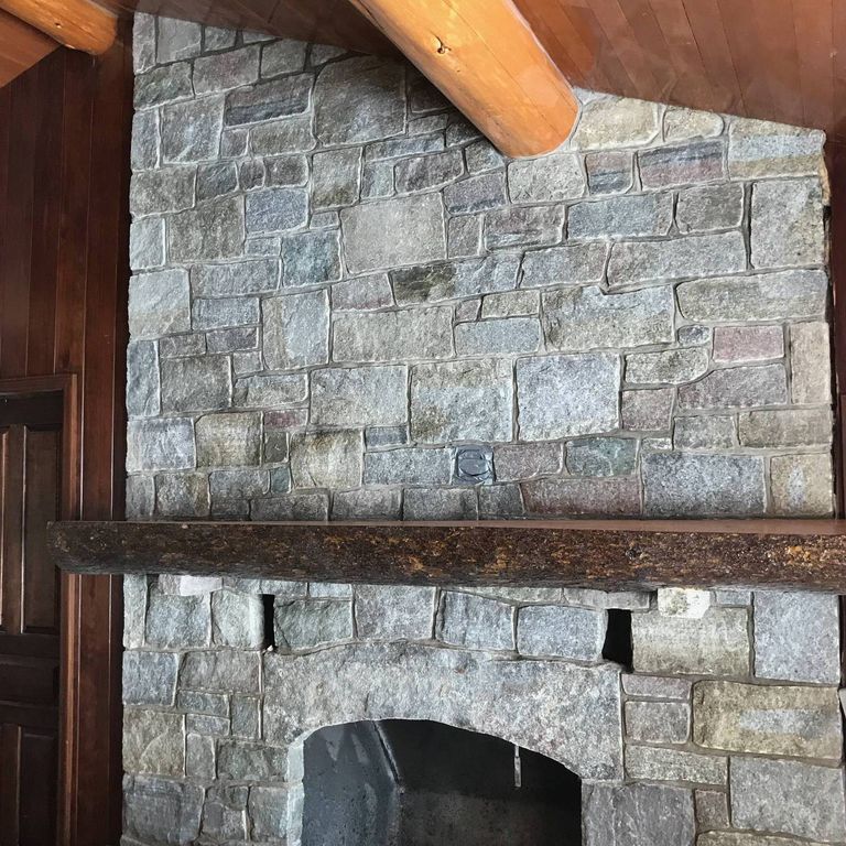 Application image for Granite - Adirondack Elite. The image shows this material being used for fireplace surround, full bed veneer, thin veneer and interior cladding. The surface finish shown is split-face with flat plane. The dominant color shown is gray; with beige, green and white secondaries. The color mix creates a cool feeling. Regular, fine speckled and random color patterns.