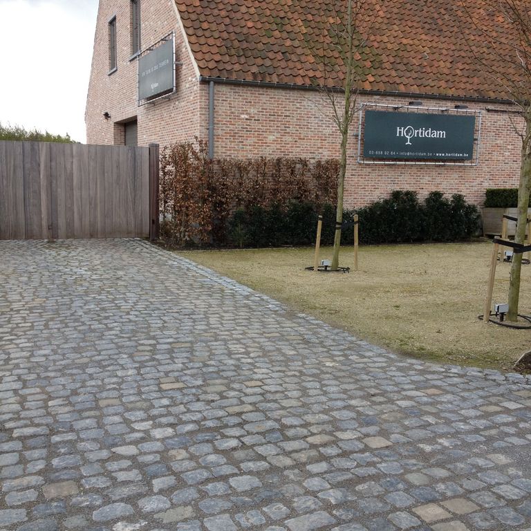 Feature image for Reclaimed Belgian Bluestone Cobblestones. The image shows this material being used in a driveway pavers application. The surface finish shown is . The dominant color shown is gray; with blue, brown, green, orange/rust, purple and tan secondaries. Speckled, blotched and cloudy color patterns.