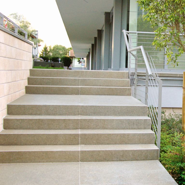 Feature image for Porphyry Worked Surface - Imperial Brown. The image shows this material being used for landings, monolithic steps and treads. Dominant colors shown are beige and gray; with brown, green, white, tan and cream secondaries. The color mix creates a warm feeling. Fine speckled color pattern.