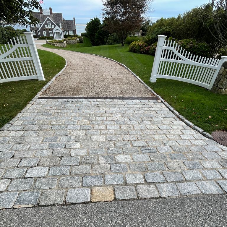 Application image for Granite Cobblestones - Gray. The image shows this material being used in a driveway aprons application. The surface finish shown is tumbled. The dominant color shown is gray; with white and tan secondaries. Exhibits 