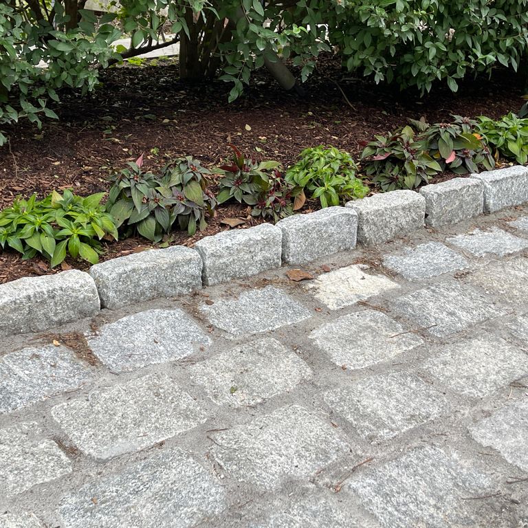 Application image for Granite Cobblestones - Gray. The image shows this material being used for landscape curbing & edging and driveway pavers. The surface finish shown is tumbled. The dominant color shown is gray; with white and cream secondaries. The color mix creates a cool feeling.