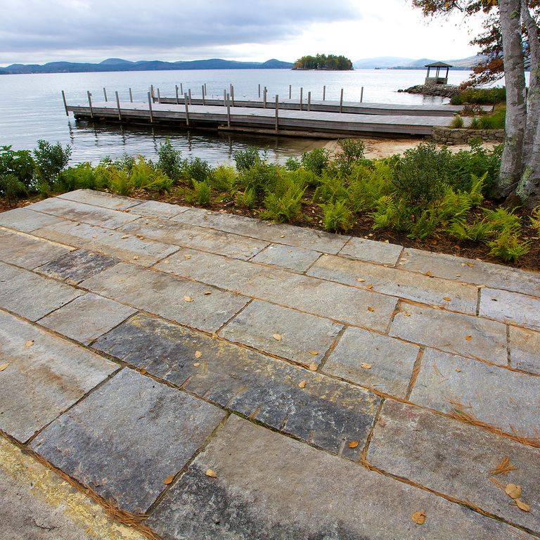 Feature image for Reclaimed First Generation Granite Curbstone. The image shows this material being used in a patio pavers application. Design patterns shown are roughly square roughly rectangular, planks with constant or same size and planks with varied heights and lengths. The dominant color shown is gray; with beige, brown, orange/rust, purple, white and tan secondaries. The color mix creates a warm feeling. Regular, speckled and blotched color patterns.