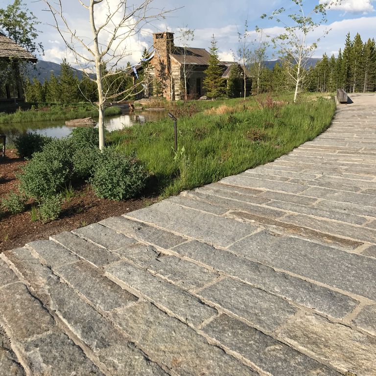Feature image for Reclaimed Second Generation Granite Curbstone. The image shows this material being used for driveway pavers and path pavers. The design pattern shown is planks with constant or same size. Dominant colors shown are brown, gray and tan; with beige, orange/rust, white and cream secondaries. The color mix creates a warm feeling. Regular, speckled, blotched and cloudy color patterns.