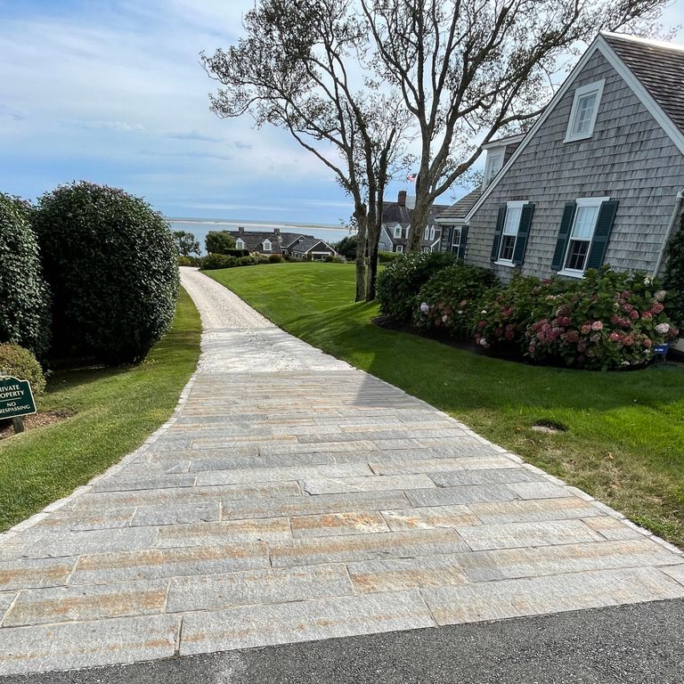 Application image for Reclaimed Third Generation Granite Curbstone. The image shows this material being used in a driveway aprons application. The surface finish shown is a combination of weathered and worn. The dominant color shown is gray; with a beige secondary. Exhibits 