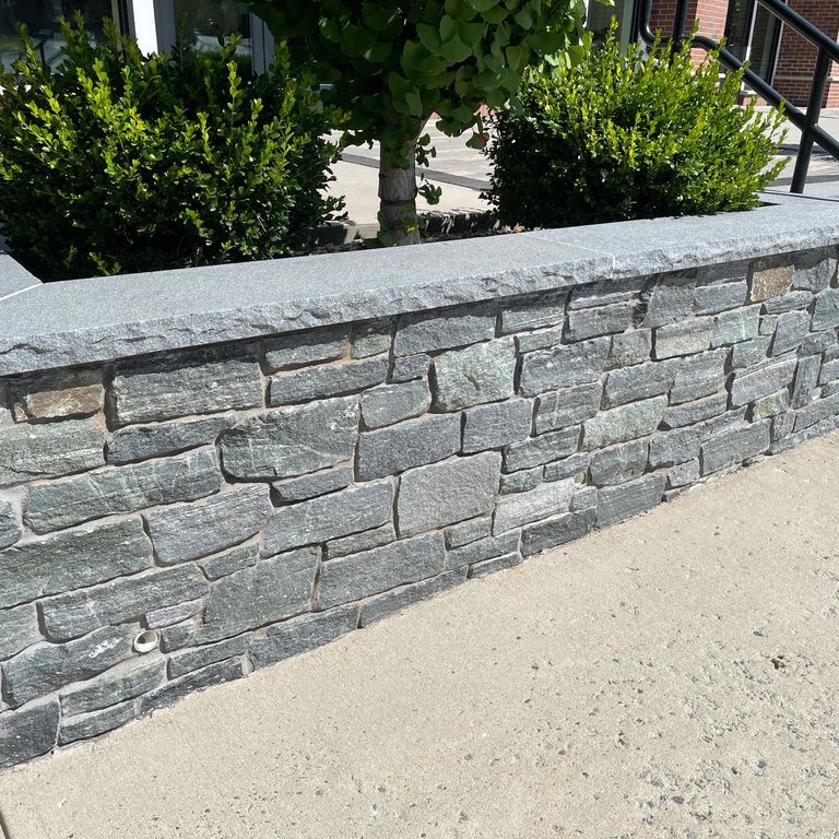 Feature image for Connecticut White Line Granite. The image shows this material being used in a landscape wallstone application. The surface finish shown is split-face. Dominant colors shown are gray and white; with green and white secondaries. Exhibits 