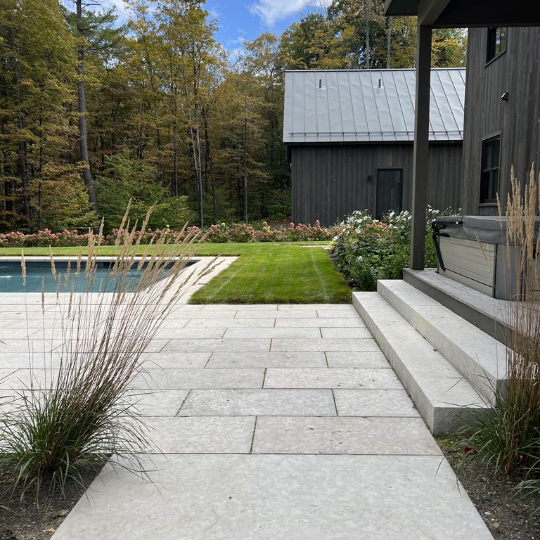 Feature image for Limestone - Madison. The image shows this material being used for pool coping, path pavers, patio pavers and monolithic steps. The surface finish shown is flamed. The dominant color shown is gray; with beige, blue, purple, white and cream secondaries. The color mix creates a cool feeling. Regular and cloudy color patterns.