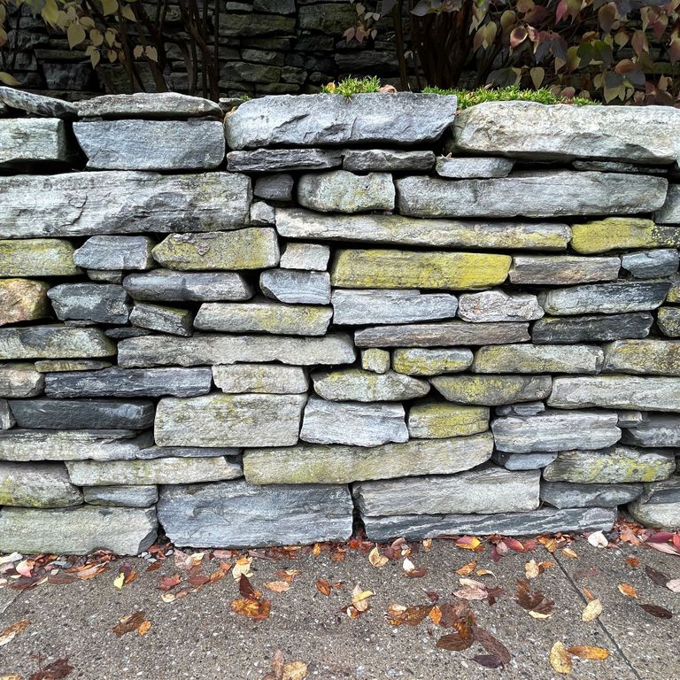 Application image for Mystic Mountain Natural - Wallstone. The image shows this product being used in a landscape wallstone application. The surface finish shown is split-face. The dominant color shown is gray; with blue, brown, green and white secondaries.