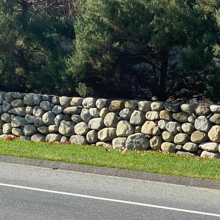 Application image for New England Fieldstone - Buff Rounds Wallstone. The image shows this product being used in a landscape wallstone application. The surface finish shown is weathered. The dominant color shown is gray.