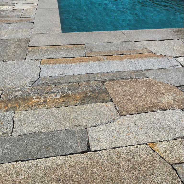 Application image for Reclaimed Second Generation Granite Curbstone Plank Pavers. The image shows this product being used in a pool deck pavers application. The surface finish shown is a combination of split-face with flat plane, weathered and worn. The dominant color shown is gray; with brown, orange/rust and tan secondaries.