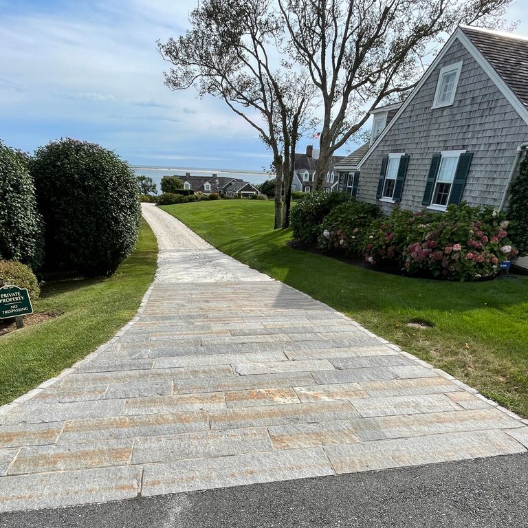 Application image for Reclaimed Third Generation Granite Curbstone Plank Pavers. The image shows this product being used in a driveway aprons application. The surface finish shown is a combination of split-face, weathered and worn. The dominant color shown is gray; with brown and orange/rust secondaries. The color mix creates a cool feeling. Regular and linear color patterns.