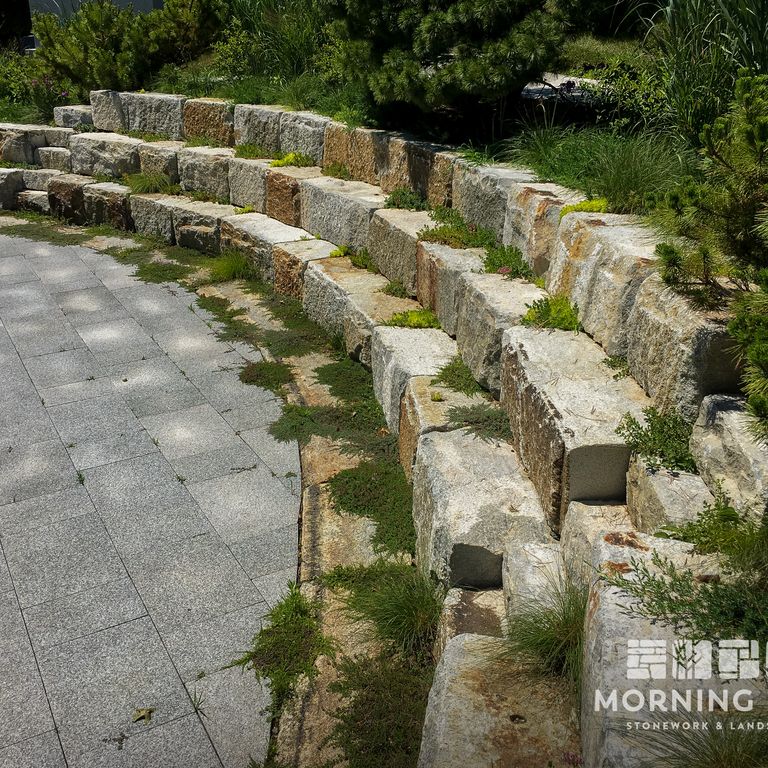 Application image for Granite - Reclaimed Block - Benches. The image shows this product being used for seating & benches and retaining wall. The surface finish shown is weathered. Dominant colors shown are brown, gray and tan. The color mix creates a warm feeling. Regular color pattern.