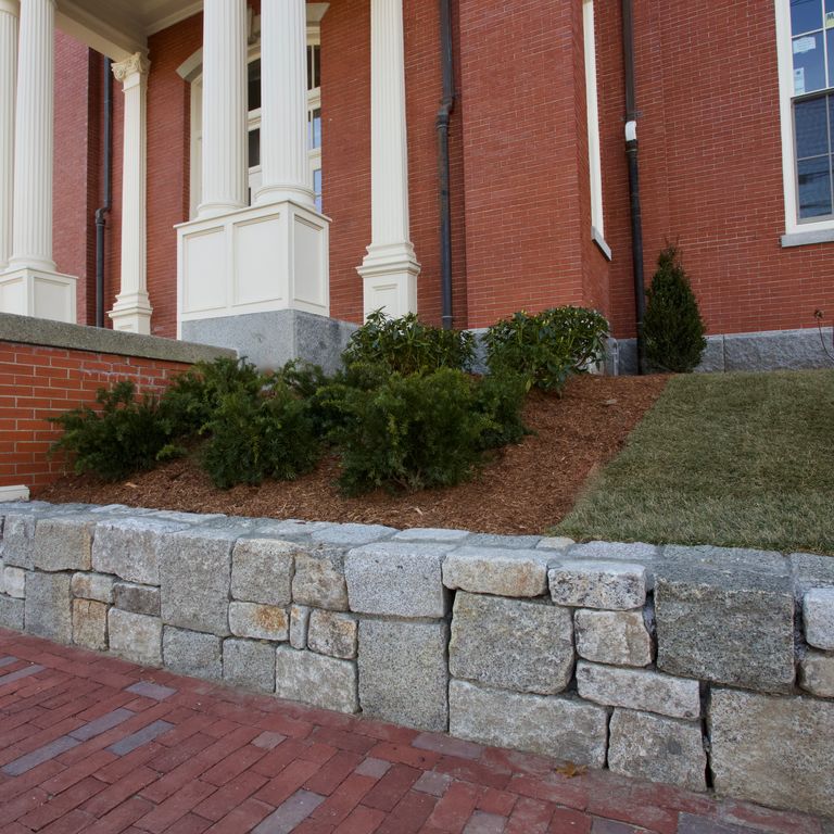 Feature image for Reclaimed Granite Block Wallstone - Split Block Mix. The image shows this product being used in a landscape wallstone application. Design patterns shown are dimensional squares & rectangles and quarried-snapped roughly square roughly rectangular with sharp edges. The dominant color shown is gray; with brown, orange/rust, purple, white and tan secondaries. Speckled and blotched color patterns.