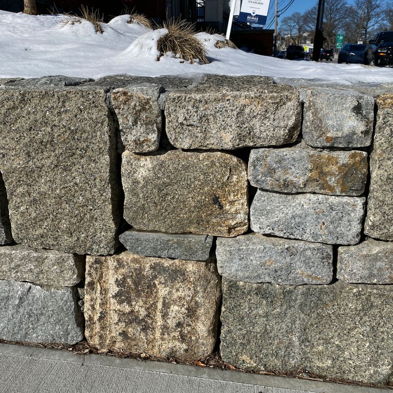 Application image for Reclaimed Granite Block Wallstone - Split Block Mix. The image shows this product being used in a landscape wallstone application. The surface finish shown is a combination of split-face and weathered. The dominant color shown is gray; with beige, brown, green and tan secondaries. The color mix creates a warm feeling. Speckled color pattern.