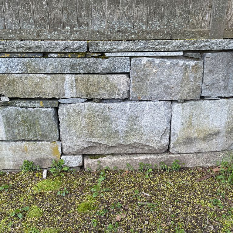 Application image for Reclaimed Granite Block Wallstone - Split Block Mix. The image shows this product being used in a landscape wallstone application. The surface finish shown is a combination of split-face and weathered. The dominant color shown is gray; with blue, brown, green and white secondaries. The color mix creates a cool feeling.
