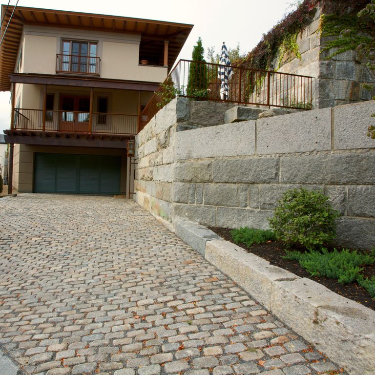 Feature image for Reclaimed New England Granite Cobblestones - Medium Pavers. The image shows this product being used in a driveway pavers application. The dominant color shown is gray; with beige, brown, green, orange/rust, purple, white, tan and cream secondaries. The color mix creates a warm feeling. Regular, speckled and blotched color patterns.