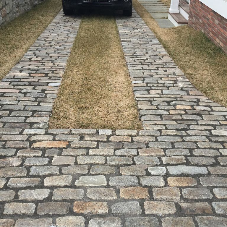 Application image for Reclaimed New England Granite Cobblestones - Medium Pavers. The image shows this product being used in a driveway pavers application. The surface finish shown is a combination of weathered and worn. The dominant color shown is gray; with beige, brown, green, orange/rust, purple, white, tan and cream secondaries. The color mix creates a warm feeling. Fine speckled and speckled color patterns.