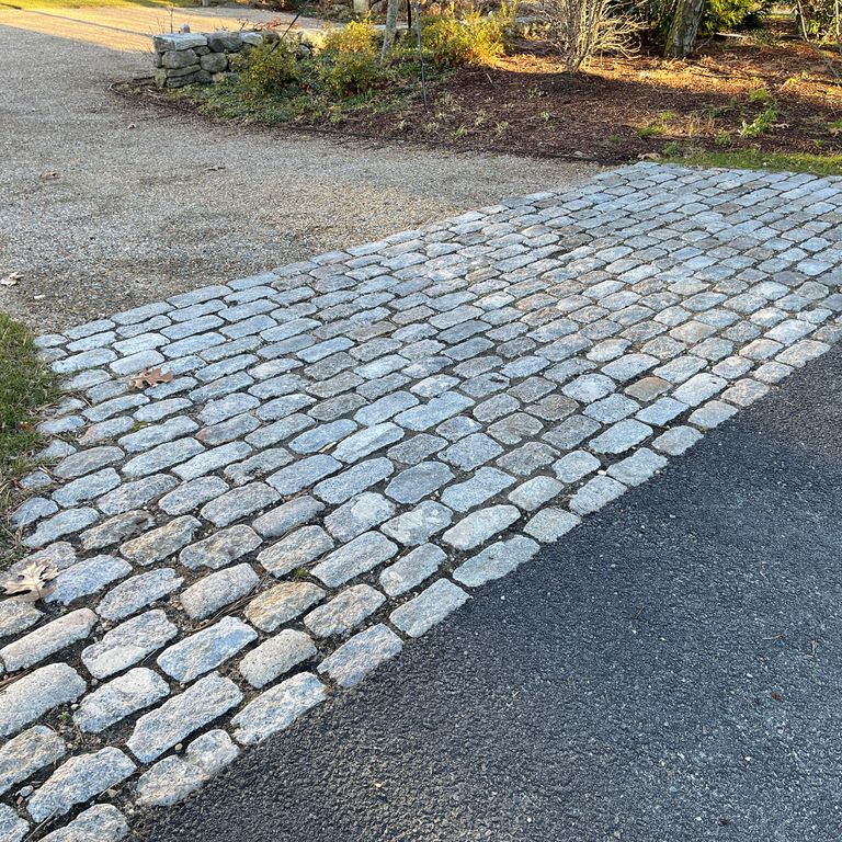 Application image for Reclaimed New England Granite Cobblestones - Medium Pavers. The image shows this product being used in a driveway pavers application. The surface finish shown is a combination of weathered and worn. The dominant color shown is gray; with brown and orange/rust secondaries. The color mix creates a cool feeling. Speckled color pattern.