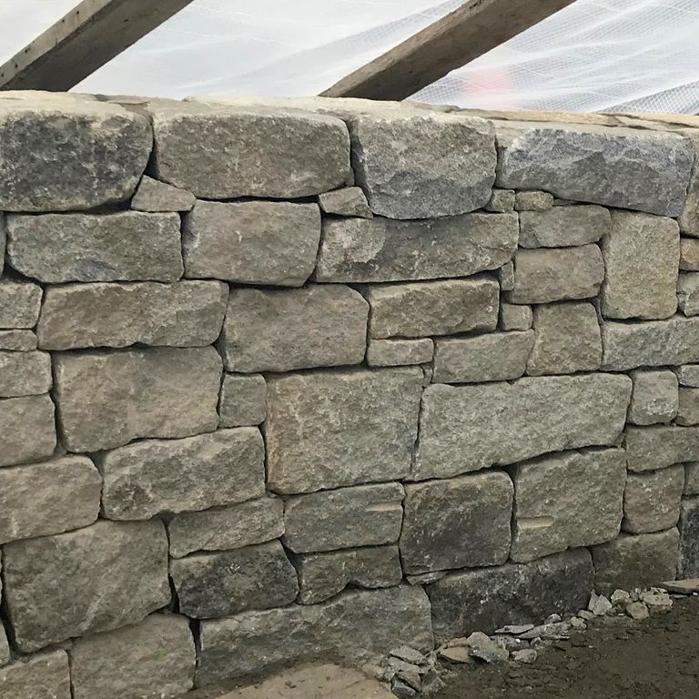 Application image for Granite - Reclaimed New England Quarry Tailings - Wallstone. The image shows this product being used in a landscape wallstone application. The surface finish shown is split-face with flat plane. Dominant colors shown are beige, gray and tan; with black, brown, green, orange/rust, white and cream secondaries. The color mix creates a warm feeling. Regular, fine speckled, swirled, speckled, blotched and cloudy color patterns.