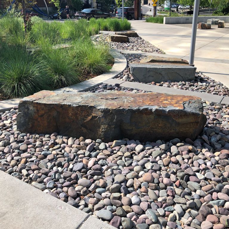 Feature image for River Rounds - Stone Mulch. The image shows this product being used in a garden accents application. The dominant color shown is gray; with beige, black, blue, brown, green, orange/rust, pink, purple, white, tan and cream secondaries. Random color pattern.