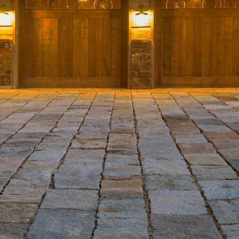 Application image for Reclaimed Granite Rustic Plank Pavers. The image shows this product being used in a driveway pavers application. The surface finish shown is a combination of split-face with flat plane and weathered. The dominant color shown is gray; with blue, brown, purple and tan secondaries. The color mix creates a cool feeling. Regular color pattern.