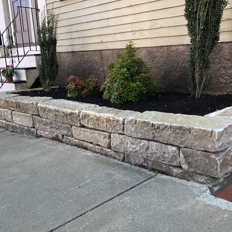 Feature image for Reclaimed Granite Curbstone - Junior Wallstone. The image shows this product being used in a landscape wallstone application. The dominant color shown is gray; with beige, brown, green, purple, white and tan secondaries. Exhibits 