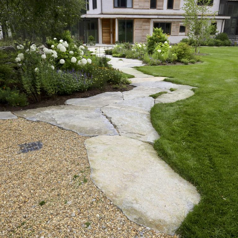 Application image for Dodlin Hill Irregular Flagstone. The image shows this product being used in a path pavers application. The surface finish shown is a combination of natural cleft and weathered. Dominant colors shown are beige, gray and cream; with white and tan secondaries. Exhibits 
