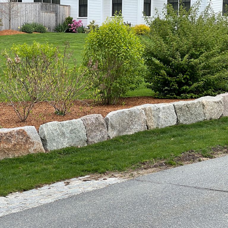 Application image for Reclaimed Granite Block - Edging. The image shows this product being used in a landscape curbing & edging application. The surface finish shown is a combination of split-face and weathered. The dominant color shown is gray; with brown, white and tan secondaries. The color mix creates a warm feeling. Speckled color pattern.