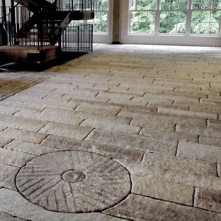 Feature image for Reclaimed Antique Mill Stones. The image shows this product being used in an interior floor application. The surface finish shown is a combination of carved, chiseled, weathered and worn. The dominant color shown is gray; with blue, brown, purple, white and tan secondaries. The color mix creates a warm feeling. Regular color pattern.