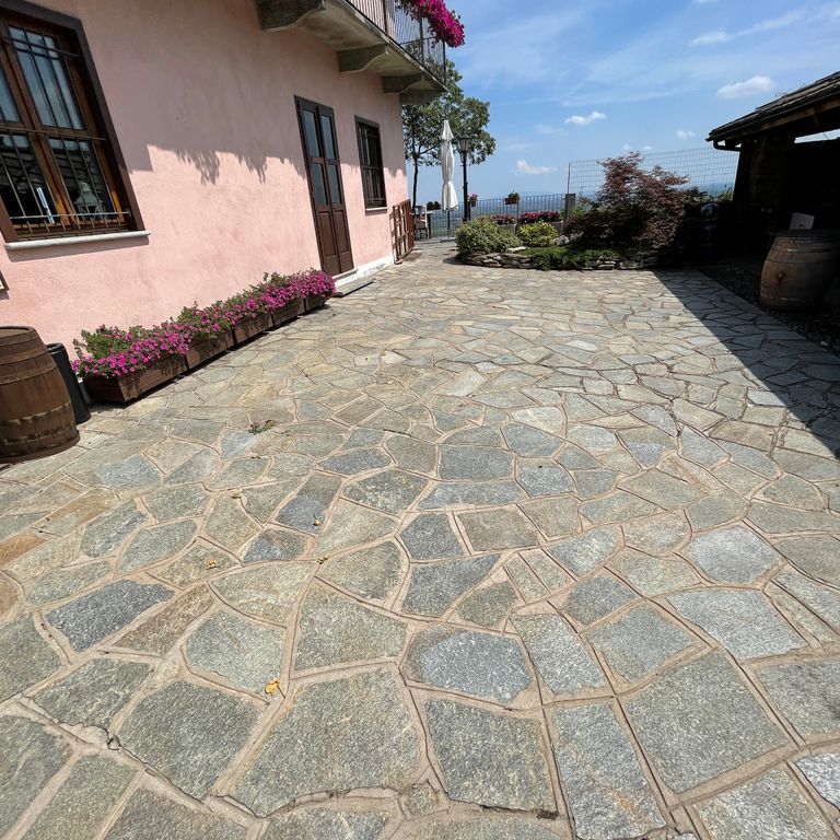 Feature image for Pietra Italia Full Color Gneiss - Irregular Pavers. The image shows this product being used in a patio pavers application. The surface finish shown is natural cleft. Dominant colors shown are beige and gray; with brown, green, orange/rust, pink, purple, white, tan and cream secondaries. The color mix creates a warm feeling. Regular, linear and random color patterns. Contains some mica and mineral inclusions.