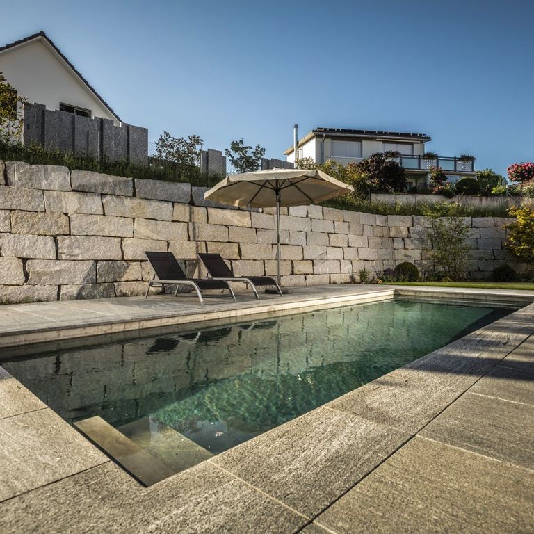 Feature image for Brazilian Lucerna Gneiss - Flamed Pavers. The image shows this product being used for pool coping, patio pavers and pool deck pavers. The surface finish shown is flamed. The dominant color shown is gray. The color mix creates a cool feeling.